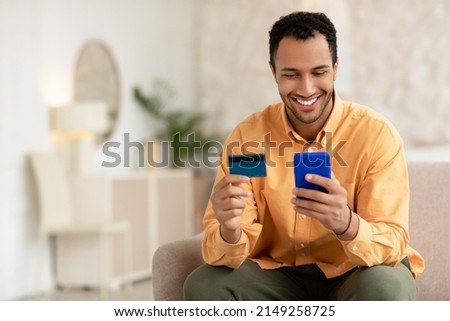 Smiling guy sitting on couch at home shopping online using smartphone holding credit card, happy young man making internet order purchase on cellphone, enter banking service system, free copy space