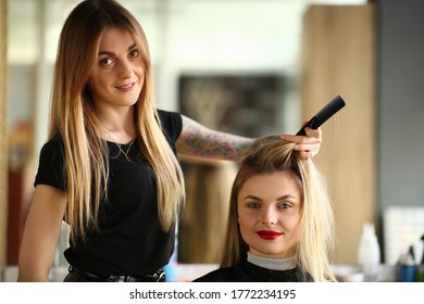 Smiling hairdresser doing hairstyle for young pretty woman sitting in a raincoat at beauty salon