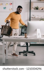 smiling handsome african american designer in orange sweater holding briefcase and looking at computer in office - Shutterstock ID 1210334296