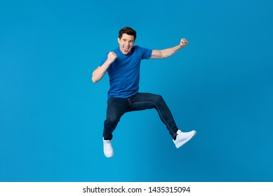Smiling handsome American man joyfully jumping and raising his fists isolated on blue studio background  fro success and freedom concepts