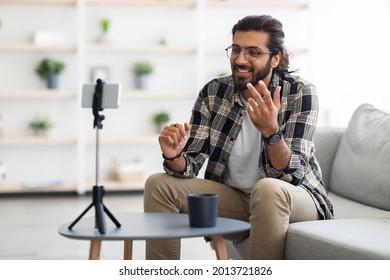 Smiling handsome arab man vlogger broadcasting from home, sitting on sofa, looking at mobile phone camera, talking, gesturing. Indian guy blogger having stream, chatting with followers, copy space