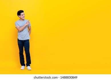 Smiling Handsome Asian Man In Plain Casual Gray T-shirt And Jeans Pointing At Empty Space Isolated On Yellow Background