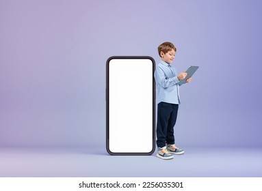 Smiling handsome boy in casual wear standing holding tablet device near large empty smartphone with mockup. Concept of child, inspired kid, social media, mobile application, education, playing game - Shutterstock ID 2256035301