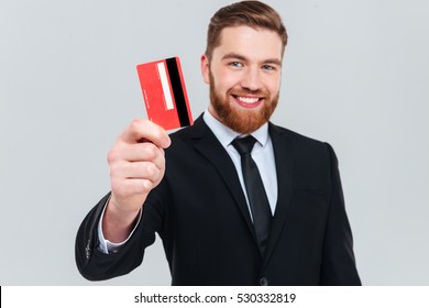 Smiling Handsome Business Man In Black Suit Holding Credit Card In Hand And Looking At Camera. Isolated Gray Background