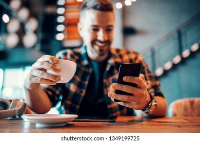 Smiling handsome Caucasian man in plaid shirt using smart phone and drinking coffee. Selective focus on hands, cafe interior. - Powered by Shutterstock