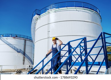 Smiling Handsome Caucasian Worker In Overall And With Helmet On Head Going Down The Stairs. Oil Production.