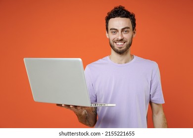 Smiling Handsome Cheerful Young Bearded Man In Casual Basic Violet T-shirt Standing Working On Laptop Pc Computer Look Camera Isolated On Orange Color Background Studio Portrait. Tattoo Translate Fun