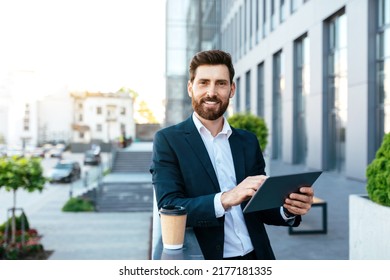 Smiling Handsome Confident European Millennial Bearded Businessman In Suit Uses Tablet To Work During Coffee Break Near Modern Office Building. Device For Business, Social Distance And Professional