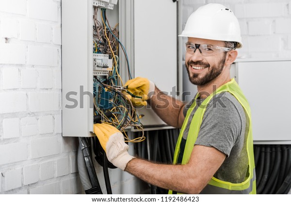 smiling handsome\
electrician repairing electrical box with pliers in corridor and\
looking at camera