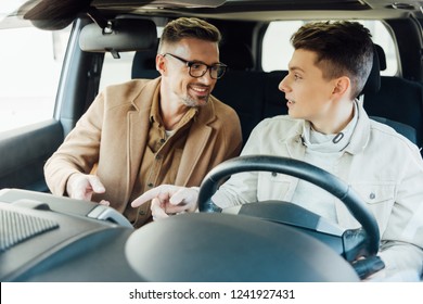 Smiling Handsome Father Teaching Teen Son Driving Car, They Looking At Each Other And Pointing On Something