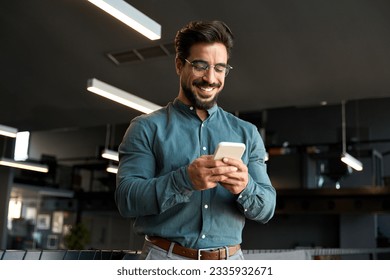 Smiling handsome Latin business man executive or employee using cell phone, happy bearded young businessman holding smartphone working on cellphone technology standing in modern office space.