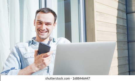 Smiling Handsome Man On A Phone Call Talking To His Family Looking Into Camera. Sucessfull Casual Businessman. Self Isolating At Home On Balcony And Using Laptop.