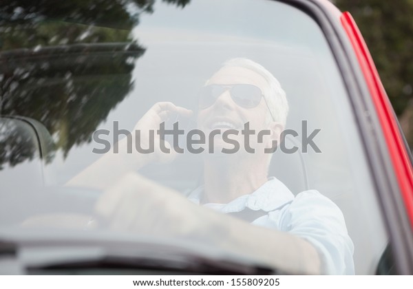 Smiling handsome man in red convertible having\
phone call while\
driving
