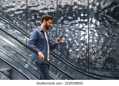Smiling handsome man using smartphone, while going down on escalator of shopping mall. Side view of bearded male in denim jacket standing on moving staircase, while messaging. Concept of lifestyle.