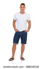 Smiling Handsome Man In White Shirt, Jeans Shorts And Black Sandals Standing With Hands In Pockets And Looking At Camera. Full Length Studio Shot Isolated On White.