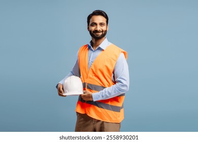 Smiling, handsome Middle aged man, happy worker wearing orange vest holding white protective helmet looking at camera standing isolated on blue background. Business concept