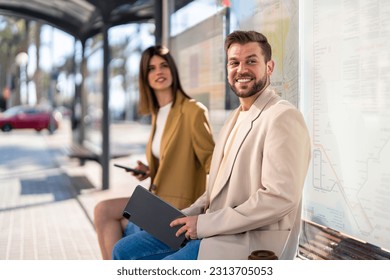 Smiling handsome millennial man with digital tablet sitting at bus station in city center with beautiful attractive young woman waiting for bus. - Powered by Shutterstock