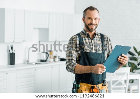smiling handsome plumber holding clipboard and looking at camera in kitchen