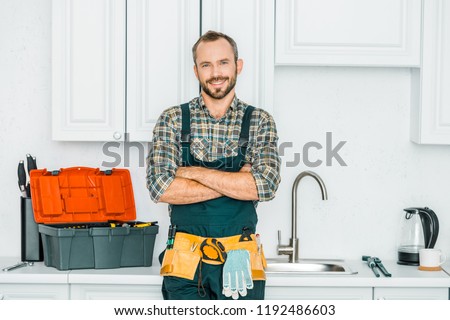smiling handsome plumber standing with crossed arms and looking at camera in kitchen