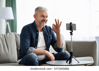 Smiling handsome senior man vlogger broadcasting from home, sitting on sofa and showing okay gesture at mobile phone camera. Male elderly blogger having stream, chatting with followers, copy space