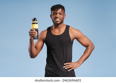 Smiling handsome young African American male athlete wearing black uniform holding water bottle looking at camera standing isolated on blue background. Concept of sports, gym, exercises - Powered by Shutterstock