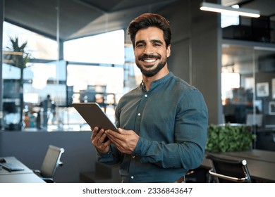 Smiling handsome young Latin business man entrepreneur using tablet standing in office at work. Happy male professional executive ceo manager holding tab computer looking at camera, portrait. - Powered by Shutterstock