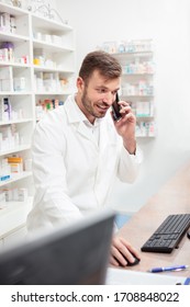 Smiling Handsome Young Male Pharmacist Talking On The Phone And Working On A Computer. Standing Behind The Counter In Pharmacy. Healthcare And Medicine Concept.