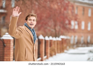 Smiling Handsome Young Man In A Trendy Coat And Scarf Waves Joyfully His Hand To Someone Walking Along Brick Houses On The City Street. Men's Fashion And Lifestyle. Copy Space.