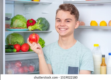 Smiling Handsome Young Teen Boy Holding Fresh Red Apple While Standing Near Open Fridge In Kitchen At Home. Portrait Of Pretty Child Choosing Food In Refrigerator Full Of Healthy Products.