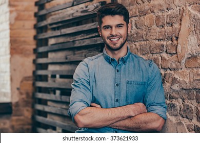 Smiling And Handsome.Handsome Young Man Keeping Arms Crossed And Looking At Camera With Smile While Standing Against Brick Wall