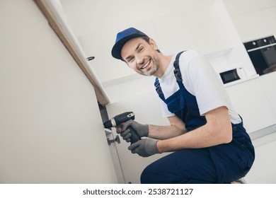 Smiling handyman in blue overalls using a cordless drill for home renovation in a modern kitchen setting - Powered by Shutterstock