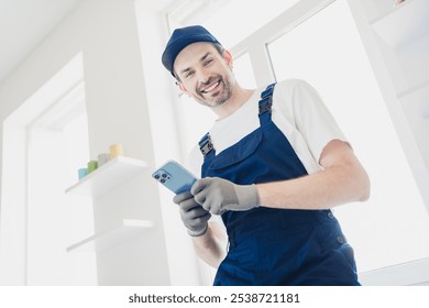 Smiling handyman in blue overalls using smartphone during home renovation in bright modern apartment - Powered by Shutterstock