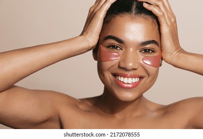 Smiling, happy african american woman with under eye patches, face mask from dark circles and puffiness, has glowing facial skin, brown background - Powered by Shutterstock