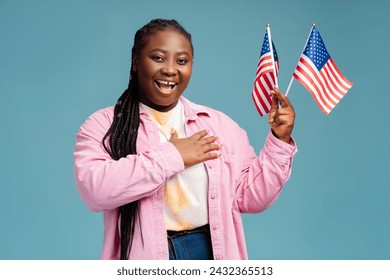 Smiling, happy African American woman holding American flag with hand on heart, looking at camera. Beautiful plus size patriot, supporter isolated on blue background. USA independence day concept - Powered by Shutterstock