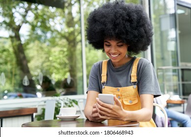 Smiling Happy African American Young Hipster Mixed Race Gen Z Young Woman With Afro Hair Holding Smart Phone, Checking Social Media, Using Mobile App Sitting At Outdoor Table In Cozy Modern Cafe.