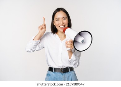 Smiling happy asian girl talking in megaphone and pointing up, announcing discount promo, showing advertisement on top, standing over white background