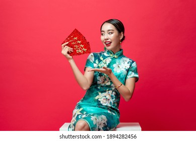 Smiling Happy Asian Woman In Oriental Cheongsam Costume Presenting Red Envelopes Or Ang Pao With Text Mean Lucky And Wealthy Isolated On Red Studio Background For Chinese New Year Celebration Concepts