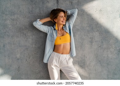 Smiling Happy Beautiful Woman Posing Against Concrete Wall On Sunny Day Wearing Denim Shirt, Yellow Top, White Pants, Summer Fashion Style Trend