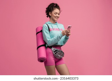 smiling happy beautiful woman in stylish sport outfit posing on pink background isolated in studio, wearing blue hoodie, bag and shorts sportswear, holding yoga mat, using smartphone - Powered by Shutterstock