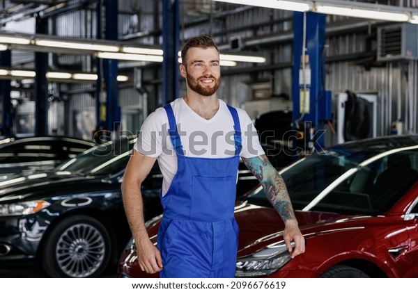 Smiling happy cheerful young male professional\
technician car mechanic repairman man in denim blue overalls white\
t-shirt walk go look aside work in light modern vehicle repair shop\
workshop indoors.