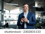Smiling happy confident mid aged male company ceo executive wearing suit holding cellphone standing in office using business mobile apps technology financial online solutions on cell phone.