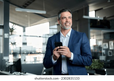 Smiling happy confident mid aged male company ceo executive wearing suit holding cellphone standing in office using business mobile apps technology financial online solutions on cell phone.