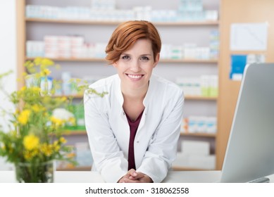 Smiling happy confident young woman pharmacist leaning on a desk in the pharmacy giving the camera a lovely big warm friendly smile