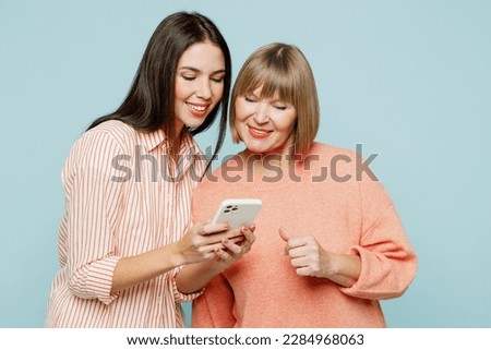 Smiling happy cool fun elder parent mom with young adult daughter two women together wear casual clothes hold in hand use mobile cell phone isolated on plain blue cyan background. Family day concept