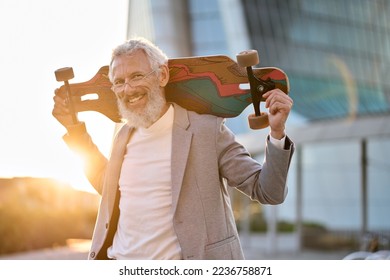 Smiling happy cool grey haired bearded older senior business man skater wearing suit holding skateboard standing in city on sunset outdoors after work. Old people freedom spirit concept. Portrait