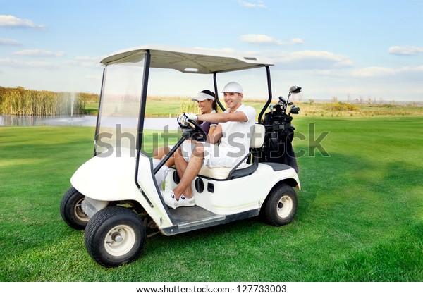 Smiling and happy couple driving a golf-cart with\
clubs on the back