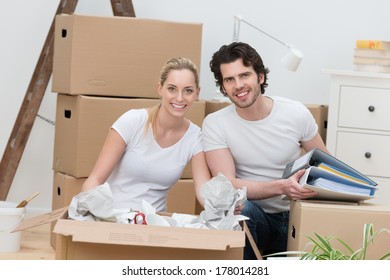 Smiling Happy Couple Unpacking Cardboard Cartons As They Sit On The Floor In Their New House Unwrapping Their Possessions And Smiling At The Camera