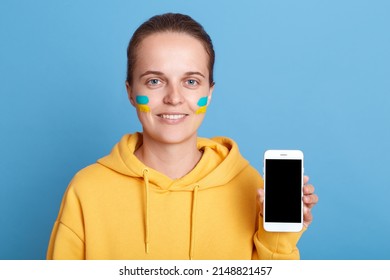 Smiling Happy Delighted Woman In Hoodie With Ukrainian Flag On Cheeks, Holding Cell Phone With Blank Screen, Showing Copy Space For Promotional Text, Posing Isolated Over Blue Background.