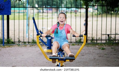 Smiling, Happy Eight Year Old Girl Engaged, Doing Exercises On Outdoor Exercise Equipment, Outdoors, In The Park, Summer, Hot Day During The Holidays. High Quality Photo