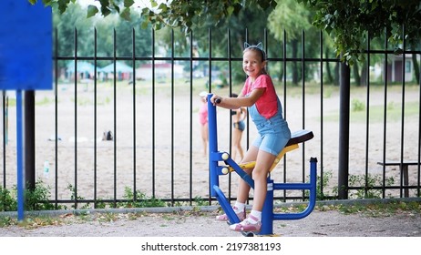 Smiling, Happy Eight Year Old Girl Engaged, Doing Exercises On Outdoor Exercise Equipment, Outdoors, In The Park, Summer, Hot Day During The Holidays. High Quality Photo